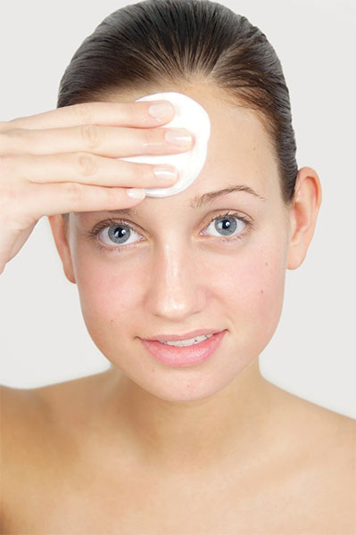 A woman with clear skin is gently wiping her forehead with a cotton pad, looking directly at the camera. She has dark hair pulled back and a slight smile, showing the importance of layering your skincare products for a radiant complexion.