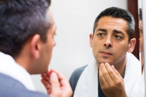A man with short dark hair looks closely at his reflection in a mirror, touching his chin with one hand. With a white towel draped over his shoulders, he appears to be inspecting his face as part of his mens skincare routine.