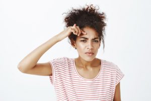 A young woman with curly hair wearing a white and red striped shirt looks confused or thoughtful, perhaps pondering 25 Facts About Adult Acne, as she touches her forehead and squints slightly against a plain white background.