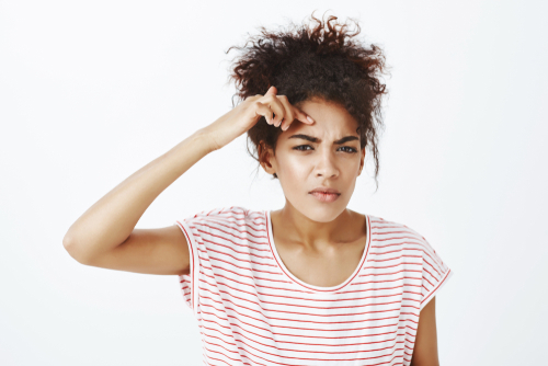 A young woman with curly hair wearing a white and red striped shirt looks confused or thoughtful, perhaps pondering 25 Facts About Adult Acne, as she touches her forehead and squints slightly against a plain white background.