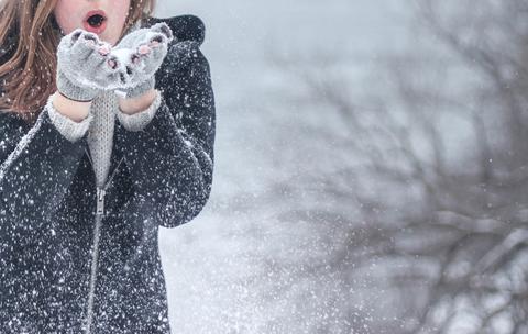 A person in a dark coat and gray gloves blows snow from their hands outdoors, with snowflakes scattering and a blurred, wintry background of bare trees.