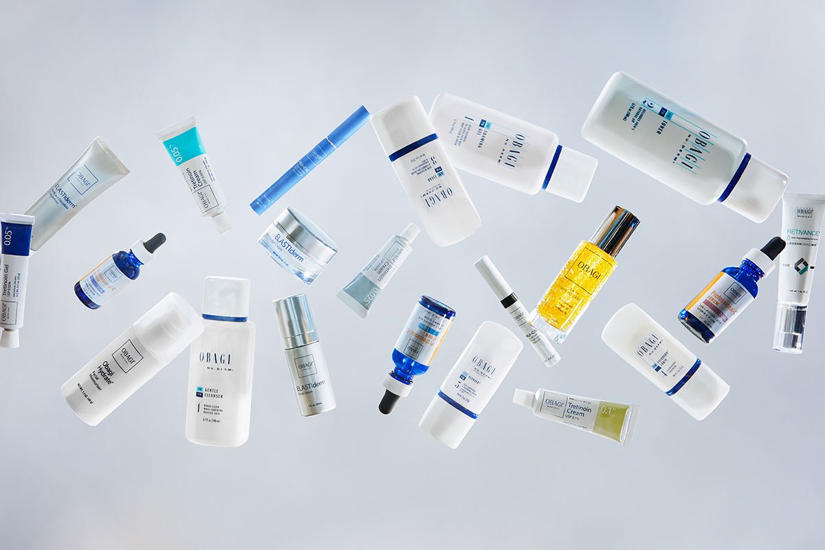 A variety of skincare product bottles and tubes are floating against a light blue background. The products include serums, creams, and lotions in white, blue, and yellow packaging, with some labeled "Obagi" and others featuring different brands.