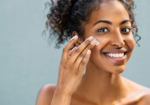 A smiling woman with curly hair applies the best Moisturizers skincare cream to her dry skin cheek with her fingertips against a light blue background. Her hair is pulled back, and she appears to be in a good mood.
