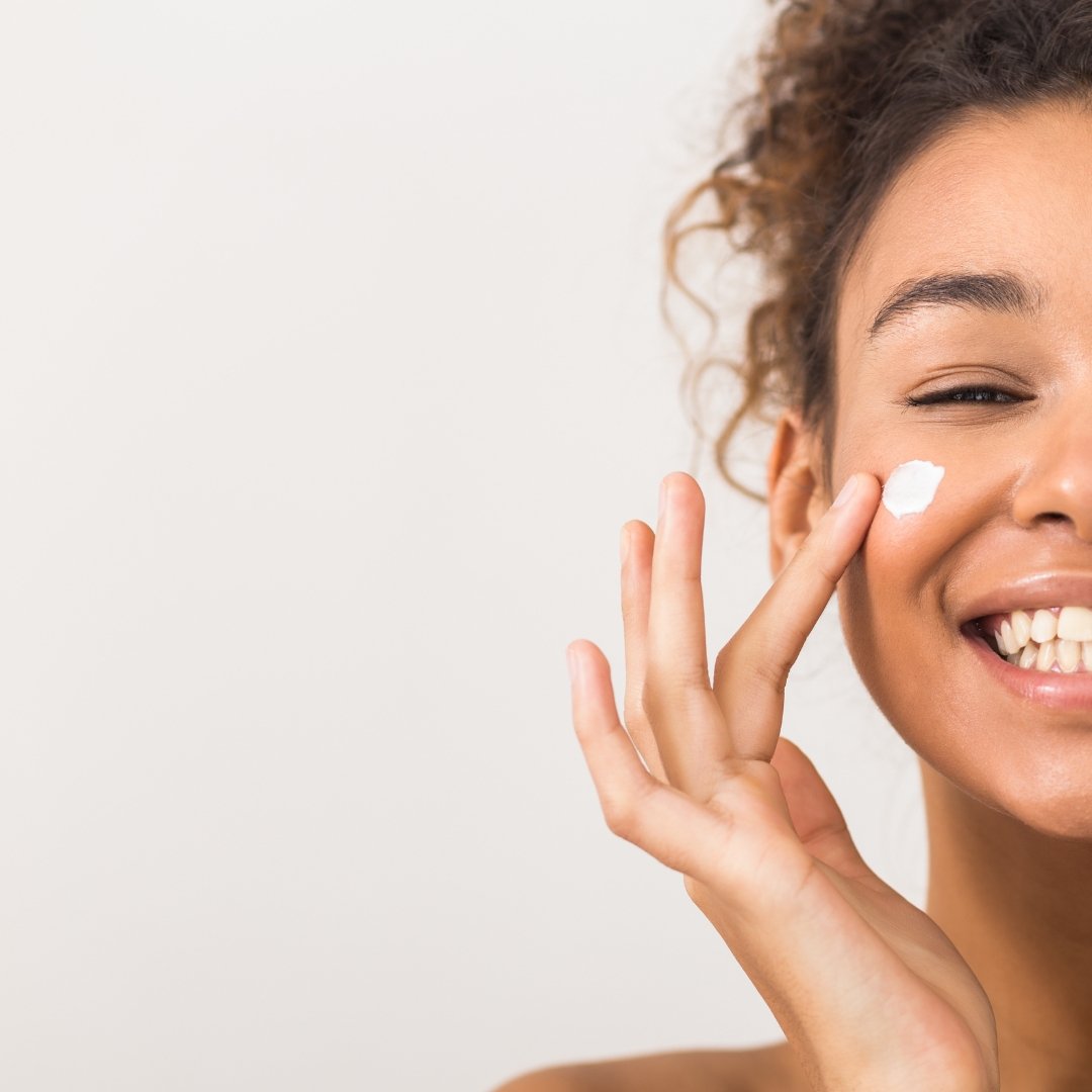 A smiling woman with curly hair applies a dab of cream to her cheek with her fingers against a plain light background.