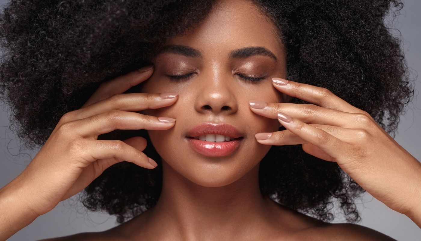 Woman with curly hair gently touching her face with eyes closed, looking relaxed and serene.