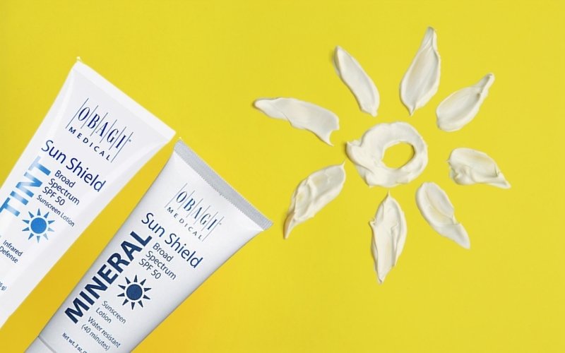 Two Obagi mineral sunscreen tubes and a sun shape made of lotion on a bright yellow background.